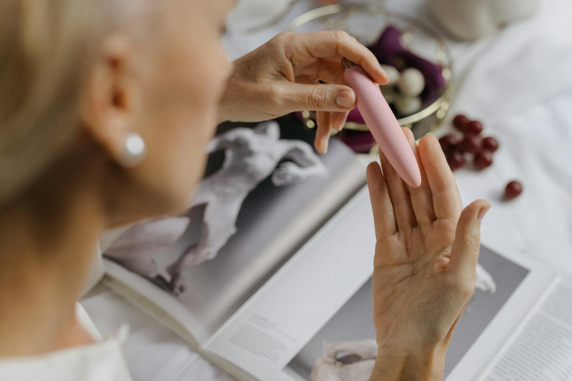 Close-up of a woman holding a pink vibrator, with art books in the background, representing intimate self-care and reconnection.