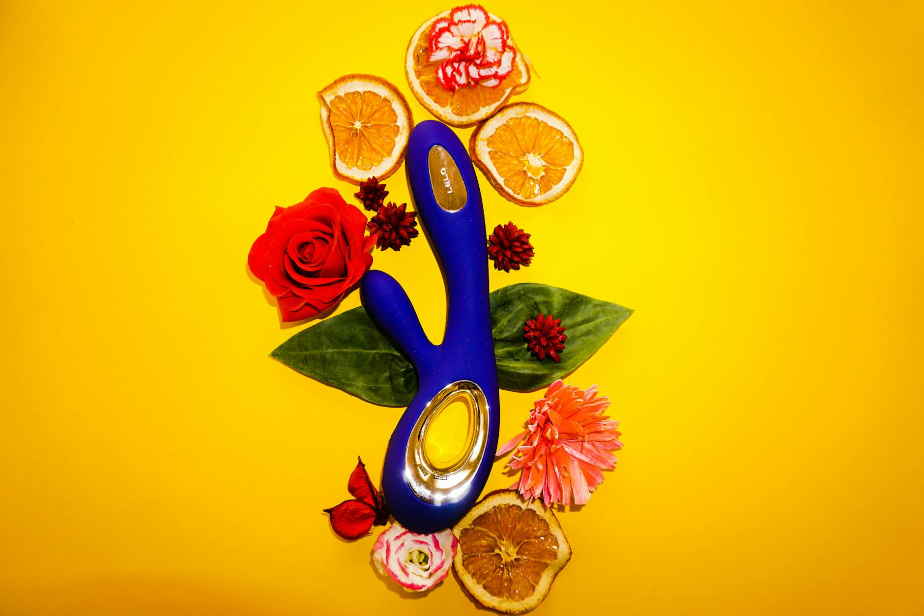 Blue lemon vibrator surrounded by flowers and citrus on a warm yellow background, symbolizing pleasure exploration
